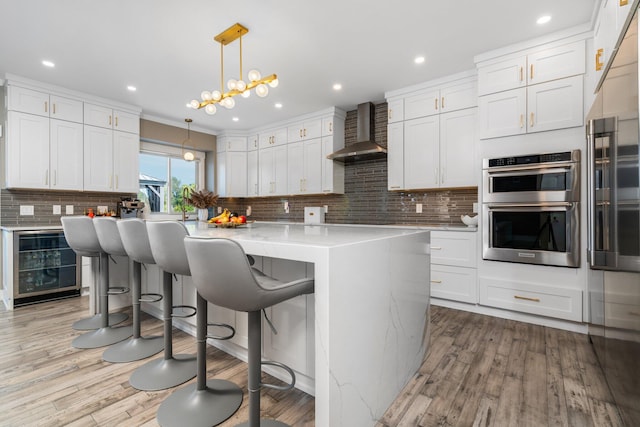 kitchen with white cabinetry, stainless steel appliances, beverage cooler, wall chimney range hood, and light wood-type flooring