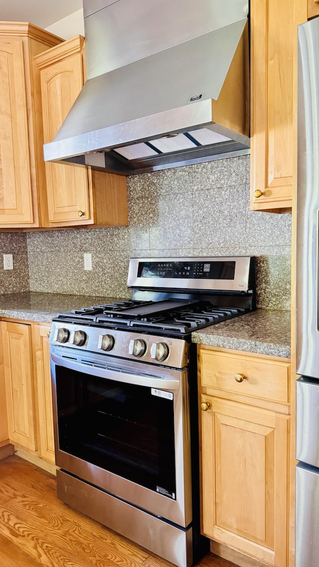 kitchen featuring wall chimney range hood, tasteful backsplash, light hardwood / wood-style flooring, light brown cabinetry, and appliances with stainless steel finishes