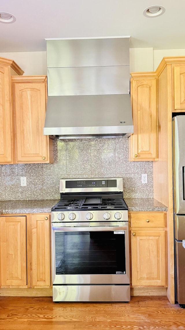 kitchen featuring light stone countertops, light brown cabinets, stainless steel appliances, wall chimney range hood, and tasteful backsplash