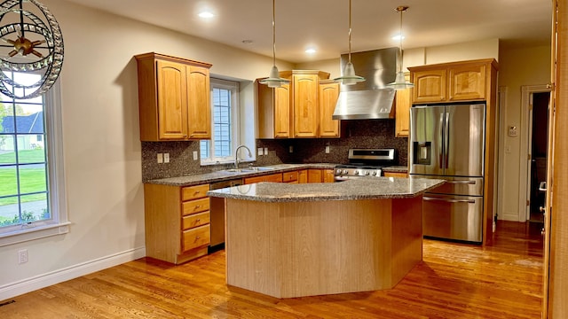 kitchen with wall chimney range hood, sink, light wood-type flooring, appliances with stainless steel finishes, and a kitchen island