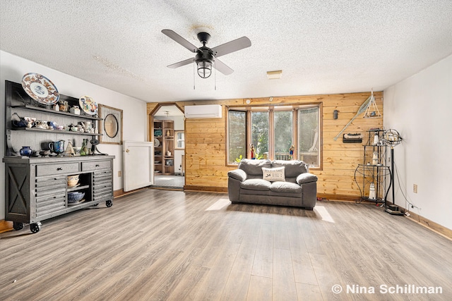 living room with a wall mounted air conditioner, wooden walls, light hardwood / wood-style flooring, ceiling fan, and a textured ceiling