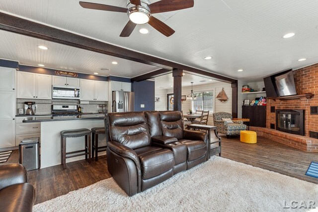 living room featuring a brick fireplace, ceiling fan, ornate columns, beamed ceiling, and dark hardwood / wood-style flooring