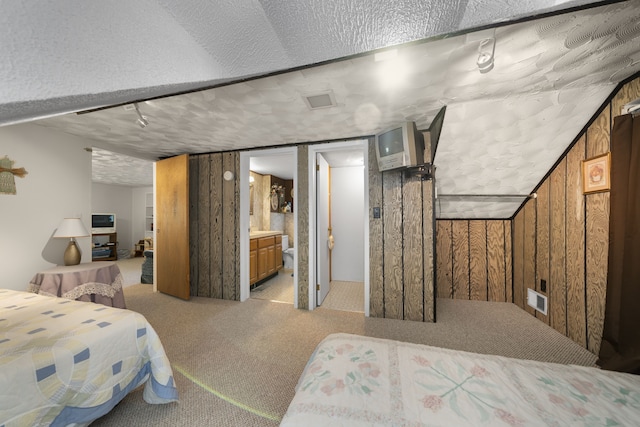 bedroom featuring light colored carpet, a textured ceiling, wooden walls, and ensuite bath