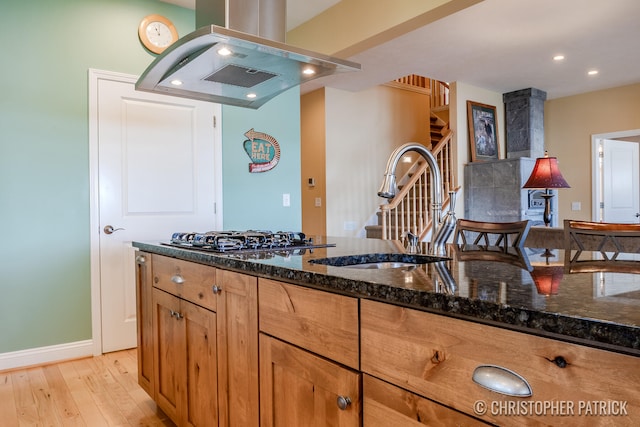 kitchen featuring stainless steel gas cooktop, a sink, light wood-style floors, dark stone countertops, and island exhaust hood