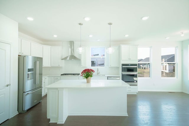 kitchen featuring stainless steel appliances, wall chimney range hood, decorative light fixtures, a center island, and white cabinetry