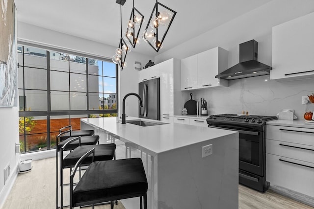 kitchen with black range, wall chimney range hood, hanging light fixtures, an island with sink, and white cabinetry