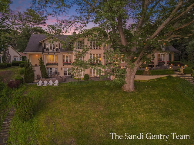 back house at dusk featuring a balcony, a patio area, and a lawn