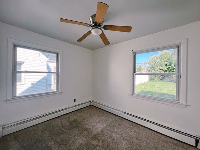 empty room featuring a baseboard heating unit, dark carpet, plenty of natural light, and a ceiling fan