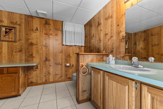 bathroom featuring a drop ceiling, vanity, wooden walls, tile patterned flooring, and toilet