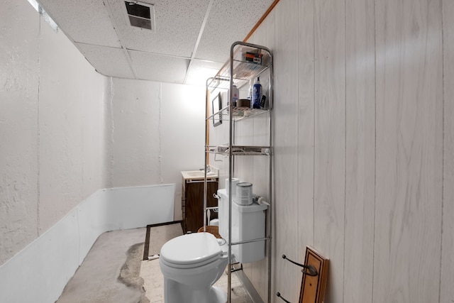 bathroom with concrete floors, toilet, a drop ceiling, and wooden walls