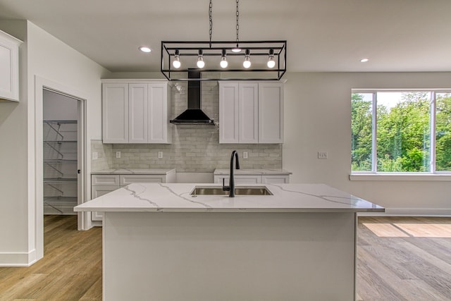 kitchen featuring white cabinets, light stone counters, a kitchen island with sink, and wall chimney range hood