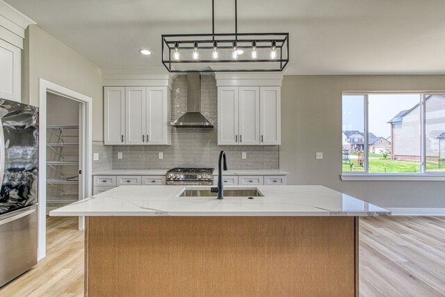 kitchen featuring light stone countertops, sink, wall chimney range hood, a center island with sink, and white cabinetry