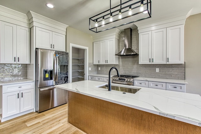 kitchen with stainless steel refrigerator with ice dispenser, a kitchen island with sink, wall chimney range hood, sink, and hanging light fixtures