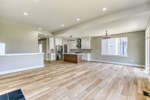 kitchen with white cabinetry, wall chimney range hood, light hardwood / wood-style flooring, an island with sink, and decorative light fixtures