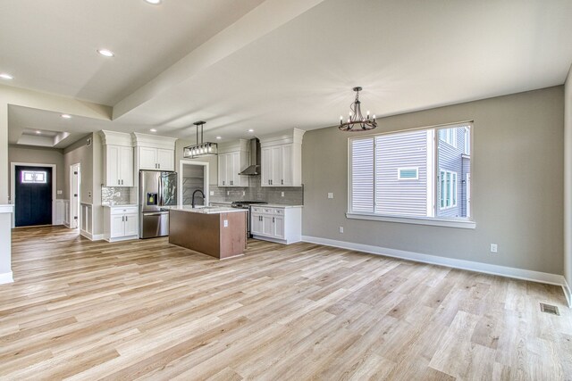 kitchen with appliances with stainless steel finishes, wall chimney exhaust hood, a center island with sink, light hardwood / wood-style floors, and hanging light fixtures