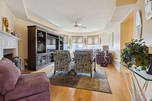 living room with ceiling fan, light hardwood / wood-style floors, a textured ceiling, a tray ceiling, and a fireplace