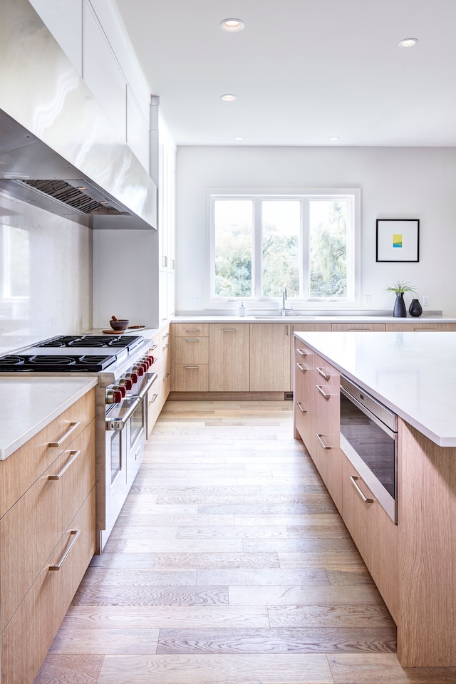 kitchen with range with two ovens, wall chimney range hood, sink, light wood-type flooring, and light brown cabinetry