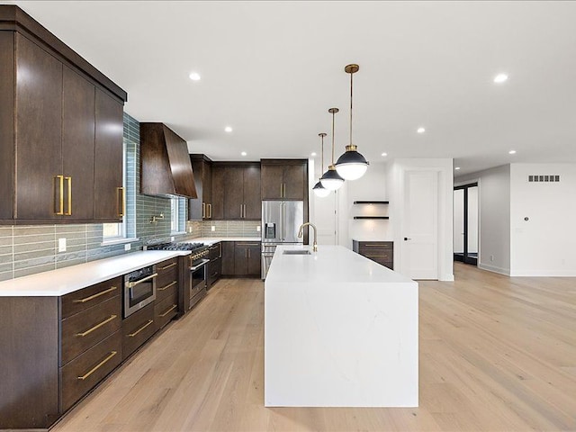 kitchen with wall chimney exhaust hood, pendant lighting, light hardwood / wood-style floors, a center island with sink, and appliances with stainless steel finishes