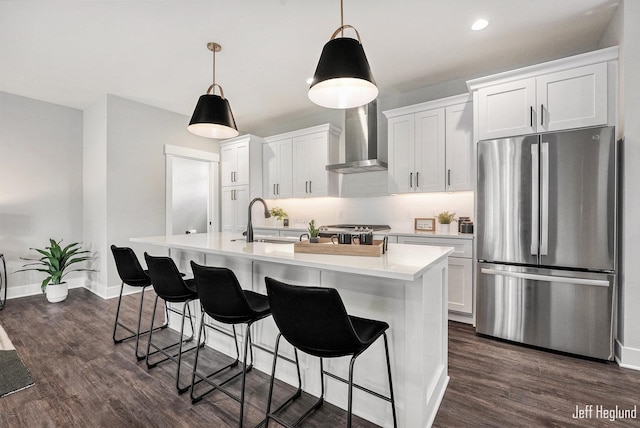 kitchen featuring white cabinets, wall chimney range hood, an island with sink, and stainless steel refrigerator