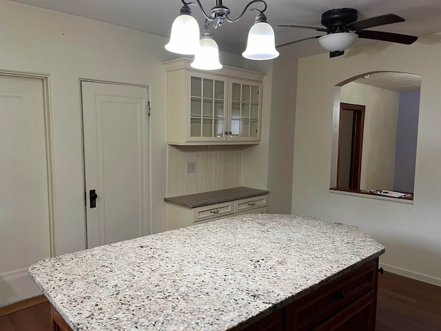 kitchen with pendant lighting, ceiling fan with notable chandelier, light stone countertops, a kitchen island, and dark hardwood / wood-style flooring