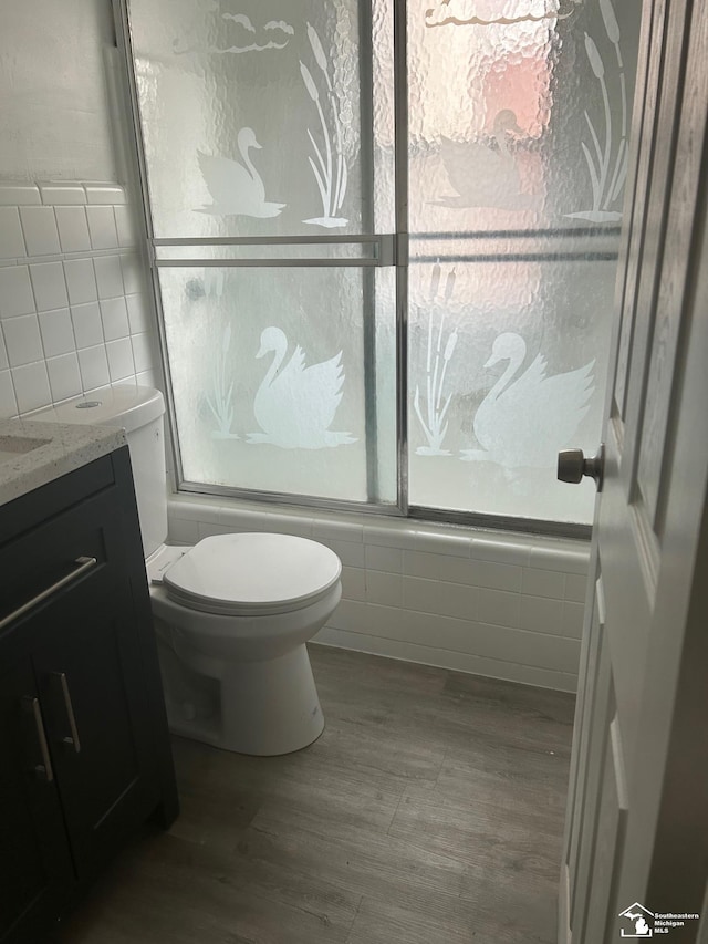 full bath with bath / shower combo with glass door, toilet, wood finished floors, vanity, and tile walls