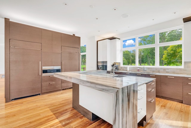 kitchen featuring a kitchen island with sink, sink, light wood-type flooring, tasteful backsplash, and light stone counters