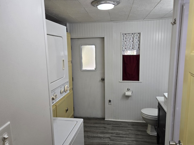 bathroom featuring stacked washing maching and dryer, wood walls, toilet, vanity, and hardwood / wood-style flooring