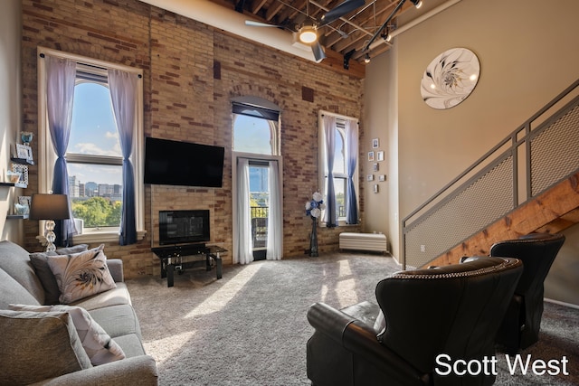 carpeted living room featuring a stone fireplace, ceiling fan, a towering ceiling, beamed ceiling, and brick wall