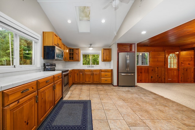 kitchen featuring ceiling fan, wood walls, light carpet, vaulted ceiling with skylight, and appliances with stainless steel finishes