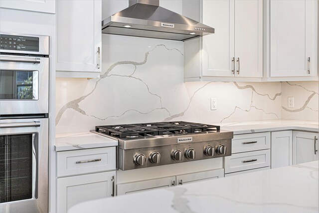 kitchen featuring light stone countertops, appliances with stainless steel finishes, tasteful backsplash, wall chimney exhaust hood, and white cabinets