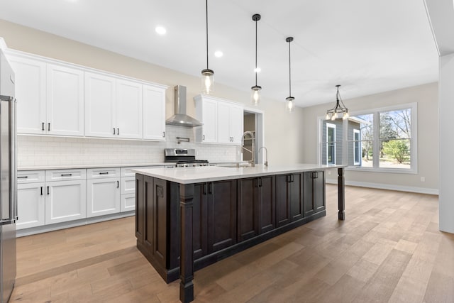 kitchen featuring wall chimney exhaust hood, stainless steel appliances, decorative light fixtures, a center island with sink, and white cabinets