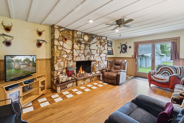 living room with wood walls, a stone fireplace, hardwood / wood-style flooring, ceiling fan, and a textured ceiling