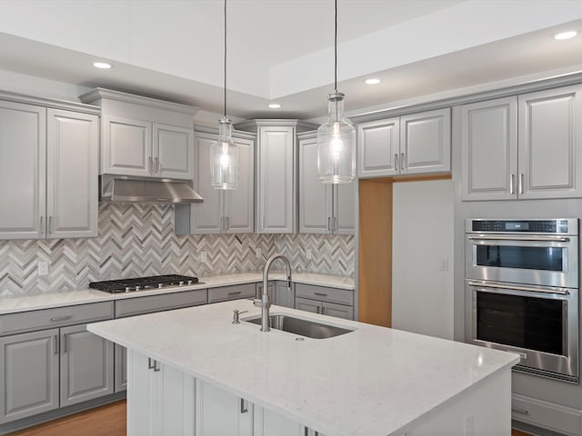 kitchen with appliances with stainless steel finishes, light stone counters, a kitchen island with sink, sink, and hanging light fixtures