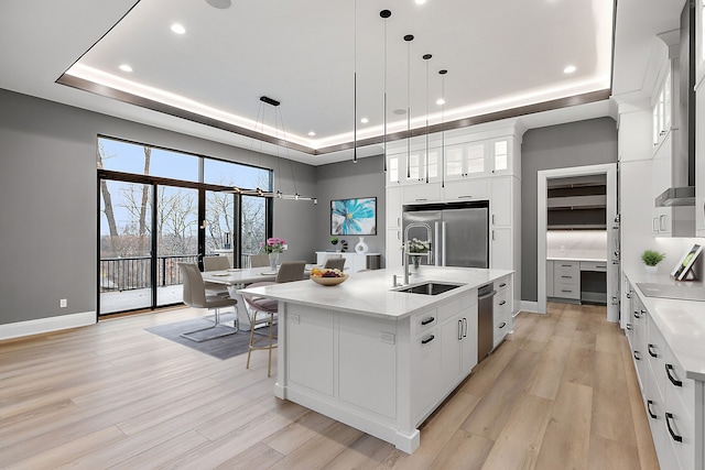 kitchen featuring light wood-type flooring, a tray ceiling, a kitchen island with sink, white cabinets, and appliances with stainless steel finishes