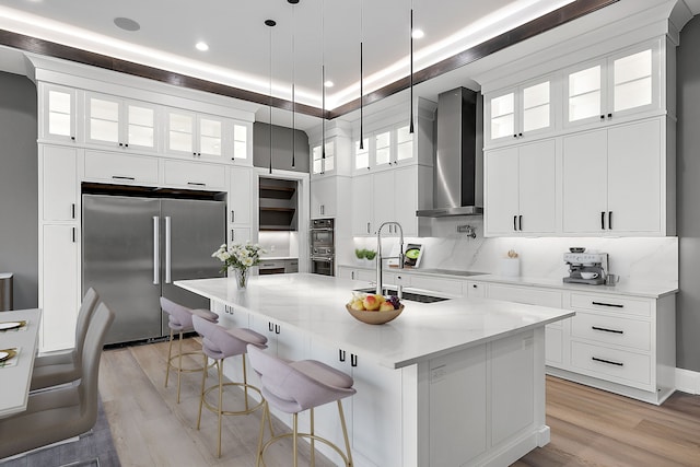 kitchen featuring black appliances, wall chimney exhaust hood, an island with sink, decorative light fixtures, and light stone counters