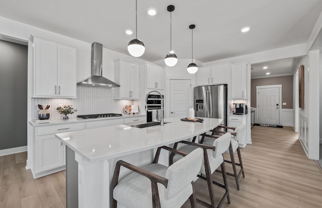 kitchen featuring appliances with stainless steel finishes, wall chimney exhaust hood, decorative light fixtures, white cabinetry, and an island with sink