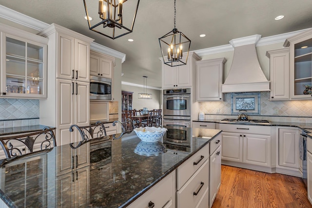 kitchen with appliances with stainless steel finishes, custom exhaust hood, light wood finished floors, glass insert cabinets, and pendant lighting