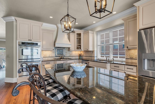 kitchen featuring stainless steel appliances, hanging light fixtures, glass insert cabinets, a sink, and premium range hood