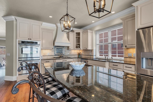 kitchen featuring appliances with stainless steel finishes, glass insert cabinets, hanging light fixtures, premium range hood, and a sink