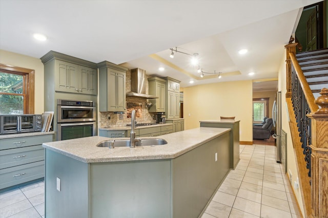 kitchen featuring sink, a kitchen island with sink, stainless steel appliances, wall chimney range hood, and decorative backsplash