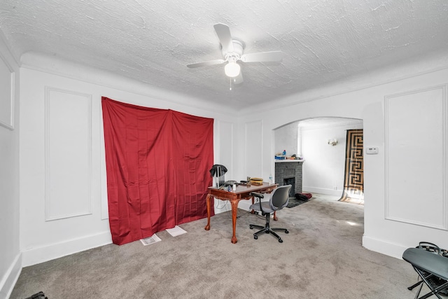 office featuring a stone fireplace, ceiling fan, light colored carpet, and a textured ceiling