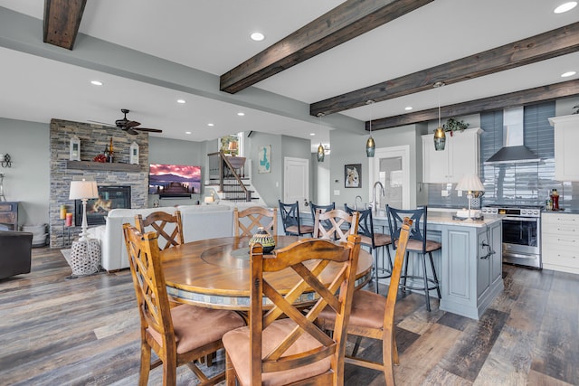 dining space with beamed ceiling, dark hardwood / wood-style floors, ceiling fan, and a stone fireplace