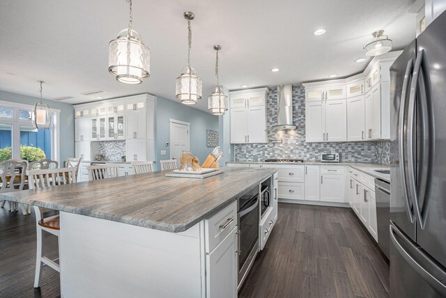 kitchen featuring appliances with stainless steel finishes, wall chimney exhaust hood, decorative light fixtures, white cabinetry, and a kitchen island