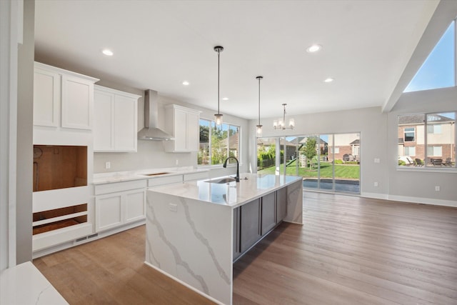 kitchen featuring a kitchen island with sink, hanging light fixtures, sink, white cabinetry, and wall chimney range hood