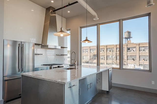 kitchen featuring a kitchen island with sink, wall chimney range hood, light stone countertops, decorative light fixtures, and stainless steel appliances