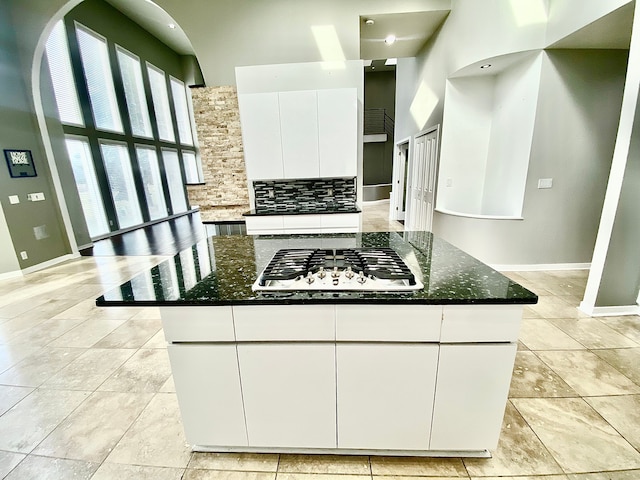 kitchen with a towering ceiling, white cabinets, decorative backsplash, dark stone counters, and stainless steel gas stovetop