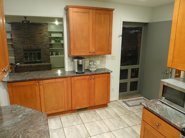 kitchen with a brick fireplace, ceiling fan, dark stone countertops, tasteful backsplash, and light tile patterned flooring