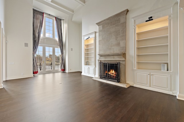 unfurnished living room with baseboards, built in features, a towering ceiling, dark wood-type flooring, and a lit fireplace