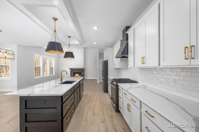 kitchen with a kitchen island with sink, stainless steel appliances, wall chimney range hood, white cabinetry, and a sink