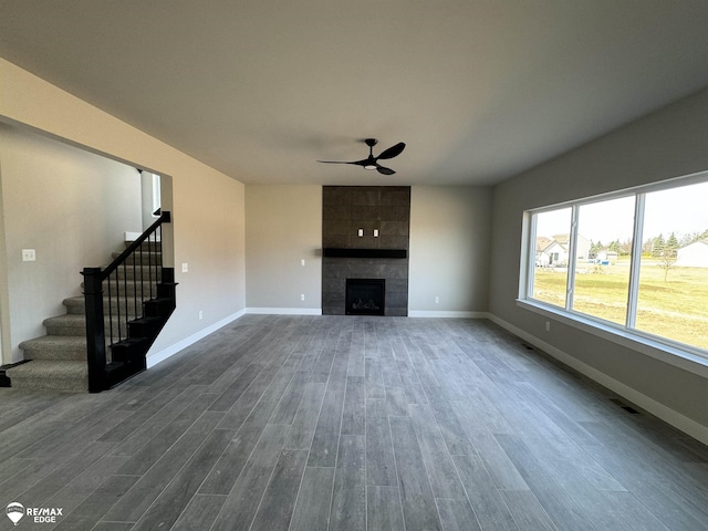 unfurnished living room featuring a tiled fireplace, dark wood-type flooring, ceiling fan, baseboards, and stairs